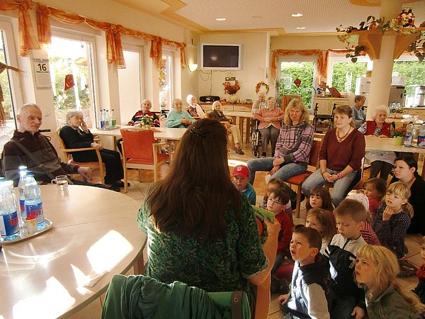 Gemeinschaftsraum mit vielen Bewohnern und Kindern. Eine Person liest den Kindern etwas vor