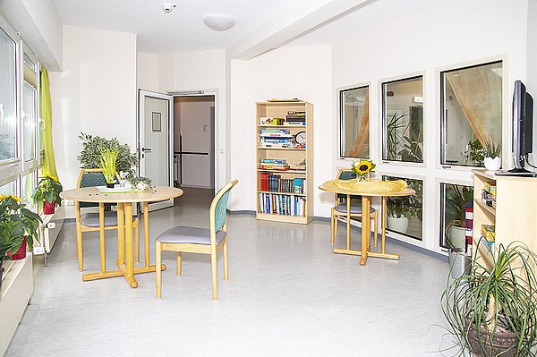 Heller Raum mit Zugang zum Wintergarten. Im Raum sind Bücherregal, Sitzgelegenheiten und ein Fernseher