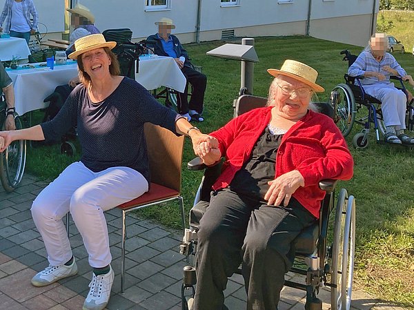 Bewohnerin in ihrem Rollstuhl im Außenbereich, freudig lachend mit einer Mitarbeiterin. Im Hintergrund sind Festtische und weitere Bewohner zu sehen.